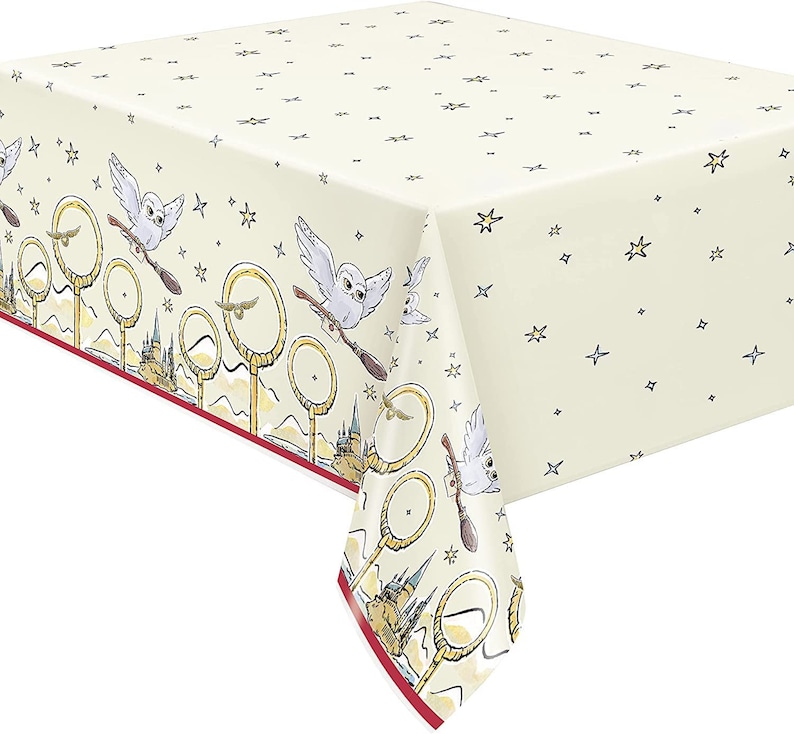 May include: Cream-colored tablecloth with a repeating pattern of stars. The border features owls on broomsticks, golden hoops, and a castle design. A red border frames the design.