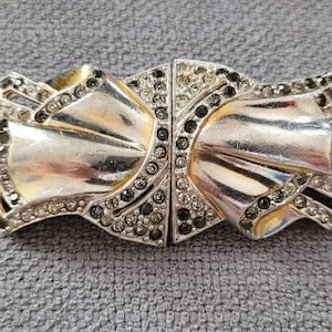 May include: A silver-toned bow-shaped clasp with a central hinge. The clasp is embellished with small, dark gray and clear rhinestones. The bow has a layered design with gold-colored accents.