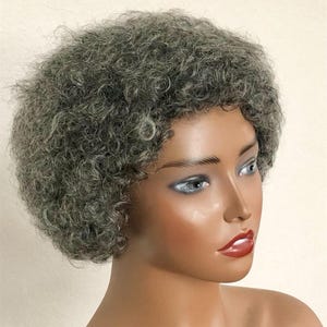 Pode incluir: Uma peruca curta e encaracolada de cor cinza sal e pimenta, estilizada num formato afro arredondado numa cabeça de manequim. O cabelo tem uma aparência texturizada e volumosa com tons de cinza misturados. O logótipo Jara Hair está impresso a preto na base.