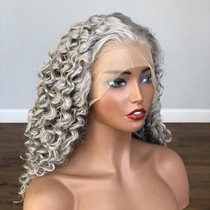 May include: A mannequin head displaying a silver-gray curly wig. The wig features a lace front and a natural-looking hairline. The mannequin has painted features, including blue eyes and red lipstick. The wig's curls are tightly wound and voluminous.