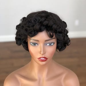 May include: A mannequin head displaying a short, curly black wig. The mannequin has painted features, including grey eyeshadow and red lipstick. The skin tone is a light tan. The background is a light grey wall and a wooden floor.