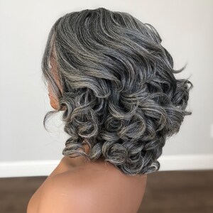K&ouml;nnte beinhalten: Graue Per&uuml;cke mit kurzem, lockigem Stil. Die Per&uuml;cke hat eine Mischung aus grauen und dunkleren Str&auml;hnen, die einen nat&uuml;rlichen Look erzeugt. Die Locken sind volumin&ouml;s und gut definiert und bieten eine klassische und elegante Frisur.