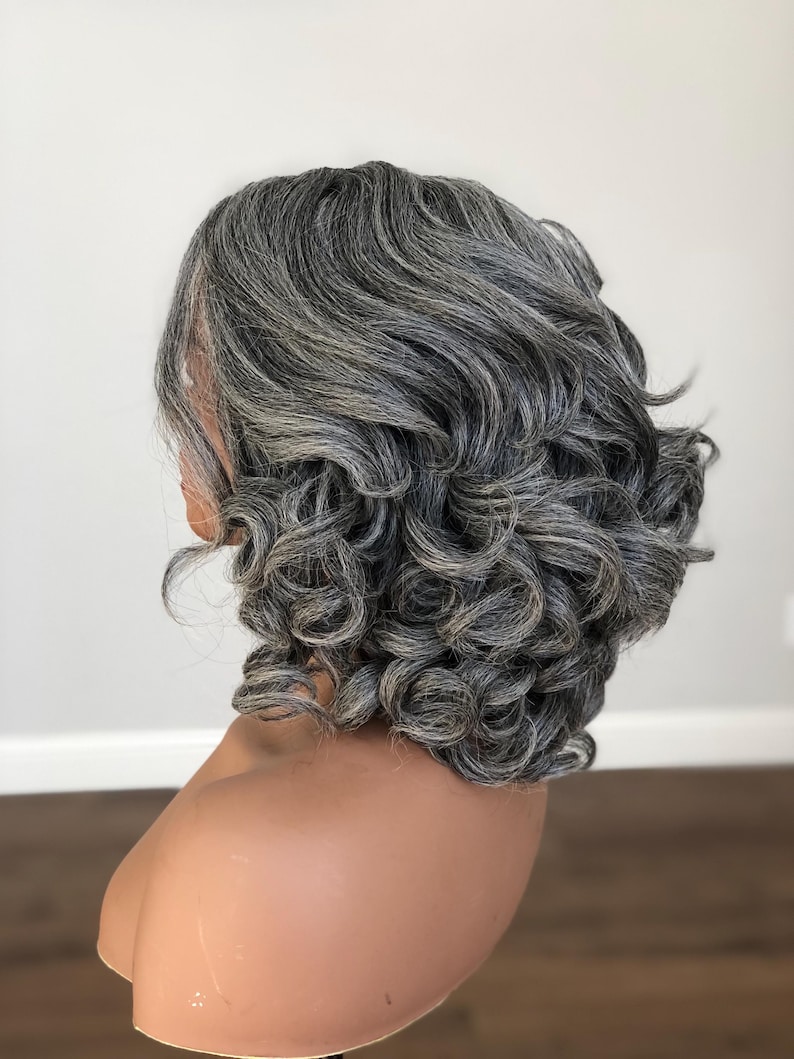 K&ouml;nnte beinhalten: Graue Per&uuml;cke mit kurzem, lockigem Stil. Die Per&uuml;cke hat eine Mischung aus grauen und dunkleren Str&auml;hnen, die einen nat&uuml;rlichen Look erzeugt. Die Locken sind volumin&ouml;s und gut definiert und bieten eine klassische und elegante Frisur.