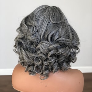K&ouml;nnte beinhalten: Eine graue Per&uuml;cke mit volumin&ouml;sem, eng gelocktem Haar. Die Per&uuml;cke ist in einer kurzen, abgerundeten Form gestylt, wobei die Locken das Gesicht umrahmen. Die Per&uuml;cke wird auf einem Schaufensterpuppenkopf pr&auml;sentiert.