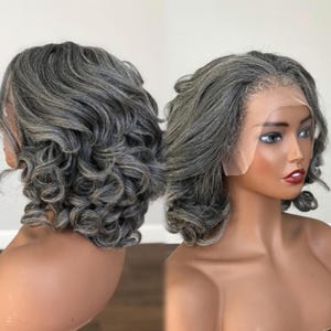 May include: A gray wig with a short, curly style. The wig features a lace front and is styled on a mannequin head. The hair is a mix of gray and silver tones, with a natural-looking hairline and soft curls.