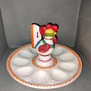 May include: A ceramic egg holder featuring a colorful rooster figurine in the center. The rooster has red, yellow, and blue accents. The white plate has eight egg-shaped indentations and an orange scalloped border.