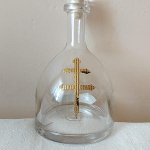 May include: A clear glass decanter with a rounded body and a clear stopper. A gold-colored cross design is centered on the front. The decanter sits on a white cloth.