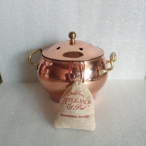 May include: A copper pot with a lid and brass handles. The lid has small holes and a brass knob. A small cloth bag with the text "APPLE JACK & Peel" hangs from the pot.