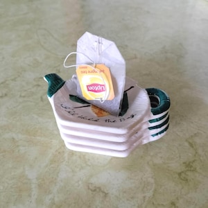 May include: A white ceramic tea bag holder shaped like a teapot, with green accents and a stackable design. A Lipton tea bag rests inside. The holder has the text "I'll hold the bag".