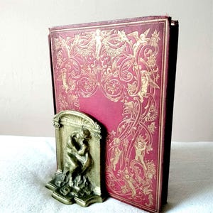 May include: A vintage book with a burgundy cover and gold embossed design, held by a bronze bookend. The bookend features a sculpted relief of two figures embracing. The book's cover has an ornate pattern with cherubs and floral elements.