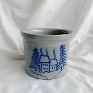 May include: A gray stoneware crock with a blue, hand-painted winter scene featuring two houses and evergreen trees. The crock has a wide, open top and a slightly textured surface. This rustic piece is perfect for kitchen decor.