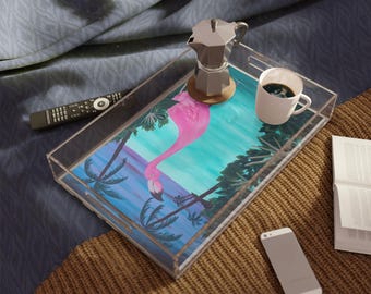 Puede incluir: Una bandeja de servicio rectangular de acrílico transparente con un diseño impreso de un flamenco rosa en un paisaje tropical con palmeras y cielo azul. La bandeja contiene una cafetera moka de metal plateado y una taza de café blanca.
