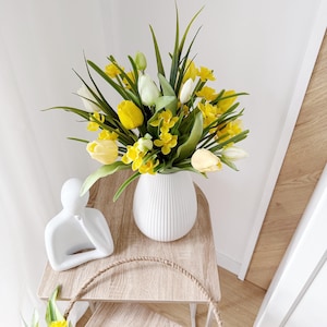 Könnte beinhalten: Eine weiße, gerippte Vase mit gelben und weißen Tulpen und Grün steht auf einem hellen Holztisch. Neben der Vase befindet sich eine kleine weiße Skulptur einer Person. Ein geflochtener Korb mit Blumen befindet sich im Vordergrund.