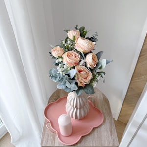 Peut inclure: Un arrangement floral avec des roses pêche et du feuillage vert dans un vase blanc cassé texturé. Le vase est posé sur un plateau rose festonné avec une bougie assortie. L'ensemble est sur une table en bois clair.
