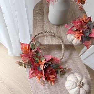 Könnte beinhalten: Ein kreisförmiger Seilkranz, verziert mit künstlichem Herbstlaub in Orange-, Rot- und gedämpften Grüntönen. Das Arrangement besteht aus verschiedenen Blättern und kleinen, stacheligen floralen Elementen, die an einer Seite des Naturfaserrings befestigt sind.