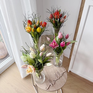 May include: Four bouquets of tulips in clear glass vases on a small, round table. The tulips are in shades of yellow, orange, red, and pink. A wooden sign that says "hello" is on the table.
