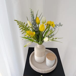 Könnte beinhalten: Ein Blumenarrangement mit gelben Tulpen, weißen Blumen und grünem Laub in einer zweifarbigen Vase. Die Vase steht auf einem beigen Tablett mit einer Kerze mit Holzdeckel. Das Arrangement steht auf einem schwarzen Tisch.
