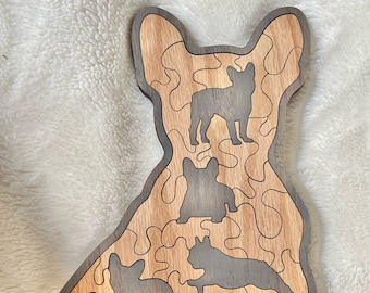 May include: A wooden puzzle featuring a silhouette of a French Bulldog with four smaller silhouettes of French Bulldogs inside. The puzzle is made of light and dark wood and has a jigsaw-like design.
