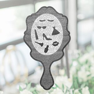 Könnte beinhalten: Ein graues Holzpuzzle in Form eines Handspiegels mit weißem Hintergrund. Das Puzzle zeigt verschiedene Bilder von Make-up, Schuhen und Modeaccessoires.