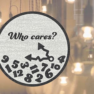 May include: A circular wooden clock with a black frame and a light grey face. The clock face displays the words "Who cares?" in black, along with black clock hands and numerals. The background is blurred with warm lighting.