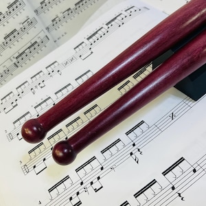 Peut inclure: Une paire de baguettes de batterie en bois violet foncé avec des embouts arrondis, placées en diagonale sur une partition de percussion imprimée présentant des motifs rythmiques et des portées pour la pratique.