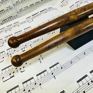 Peut inclure: Deux baguettes de batterie en bois brun foncé avec des embouts ronds reposant sur un support noir, placées sur une feuille de papier blanc imprimée de partitions de percussions et de motifs rythmiques avec des marques L et R.
