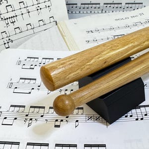Peut inclure: Deux baguettes de batterie en bois brun clair reposant sur un support noir, placées sur une partition musicale blanche avec des notations de percussion noires et le texte SOLO No. 21 imprimé sur la page.