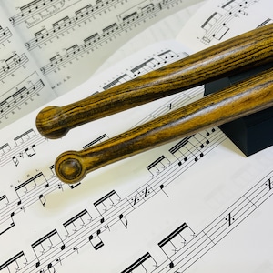 Peut inclure: Deux baguettes de batterie en bois avec un motif de grain brun foncé distinct et des embouts arrondis reposant sur une partition de musique pour percussions avec des motifs rythmiques et les lettres R et L.