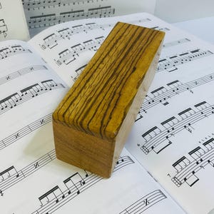 May include: A rectangular wooden percussion block featuring a distinct dark brown and tan striped grain pattern on its top surface, with smooth, light brown sides and ends, resting on a sheet of printed musical notation for percussion instruments.