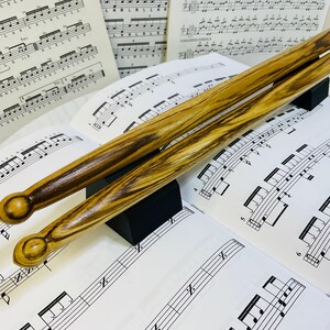 Peut inclure: Une paire de baguettes de batterie en bois avec un motif de grain brun et beige distinctif, dotées d'embouts arrondis. Les baguettes reposent sur un support rectangulaire noir, placées sur plusieurs feuilles de notation musicale imprimée.