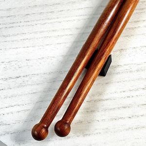 Peut inclure: Deux baguettes de batterie en bois brun poli et lisse, dotées d'embouts sphériques arrondis. Les baguettes sont placées parallèlement l'une à l'autre, révélant une texture de grain de bois riche et chaleureuse ainsi qu'une forme cylindrique.