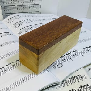 May include: A rectangular wooden box featuring a dark brown, textured lid with a distinct grain pattern and a lighter, smooth-grained tan wooden base. The box is constructed with two contrasting wood tones and sits on a sheet of printed percussion music.