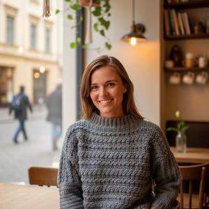 Peut inclure: Une femme porte un pull en crochet gris texturé avec un col montant. Le pull présente un motif de point détaillé. L'arrière-plan comprend un cadre de café avec des plantes suspendues, une fenêtre et des tables en bois.