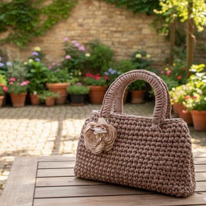 May include: A brown crocheted handbag with a flower accent. The bag has two handles and a rectangular shape. The bag is sitting on a wooden table. The background features a brick wall and potted plants.