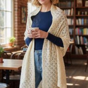 May include: A cream-coloured, open-weave shawl draped over a person wearing a navy blue top and blue jeans. The shawl has a delicate, lacy pattern. The person is holding a cup, possibly of coffee or tea, in a cafe setting.