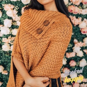 Peut inclure: Poncho en crochet jaune moutarde avec un col roulé et deux gros boutons en bois. Le poncho présente un motif de point texturé et des franges en bas. Les mots "Hooked happy" sont dans le coin inférieur droit.