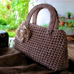 May include: A brown crocheted handbag with a flower accent. The bag features a rectangular shape with a braided handle and a decorative flower on the front. The bag is made of a thick, textured yarn.