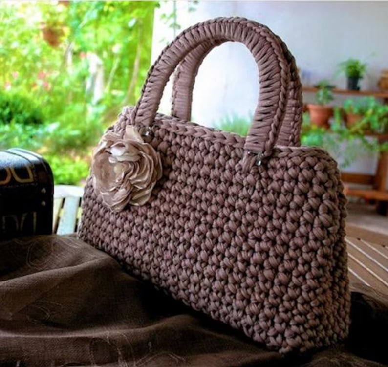 May include: A brown crocheted handbag with a flower accent. The bag features a rectangular shape with a braided handle and a decorative flower on the front. The bag is made of a thick, textured yarn.