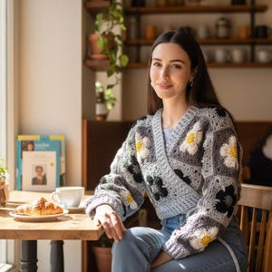 Könnte beinhalten: Eine Frau trägt eine grau-schwarz-weiße Häkeljacke mit Gänseblümchenmustern. Sie sitzt in einem Café an einem Holztisch mit einem Croissant und einer Kaffeetasse. Das Café ist hell und natürlich beleuchtet.