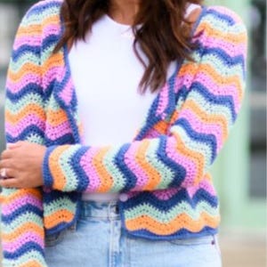Peut inclure: Un cardigan en crochet multicolore avec un motif en chevrons dans les tons de bleu, rose, orange et vert. Le pull ouvert a des manches longues et une fermeture boutonnée. Le cardigan est porté avec un haut blanc et un jean bleu.