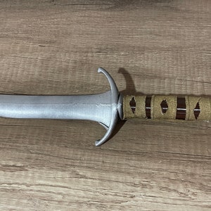 Puede incluir: Una daga de color plateado con un mango de cuero marrón. El mango tiene un patrón decorativo de pequeños círculos.