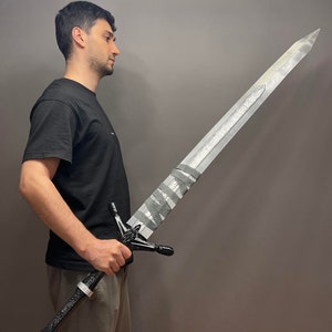 Puede incluir: Una persona sosteniendo una espada grande de color plateado con un mango negro y una empu&ntilde;adura en blanco y negro. La espada tiene un dise&ntilde;o decorativo en la hoja y una punta puntiaguda.