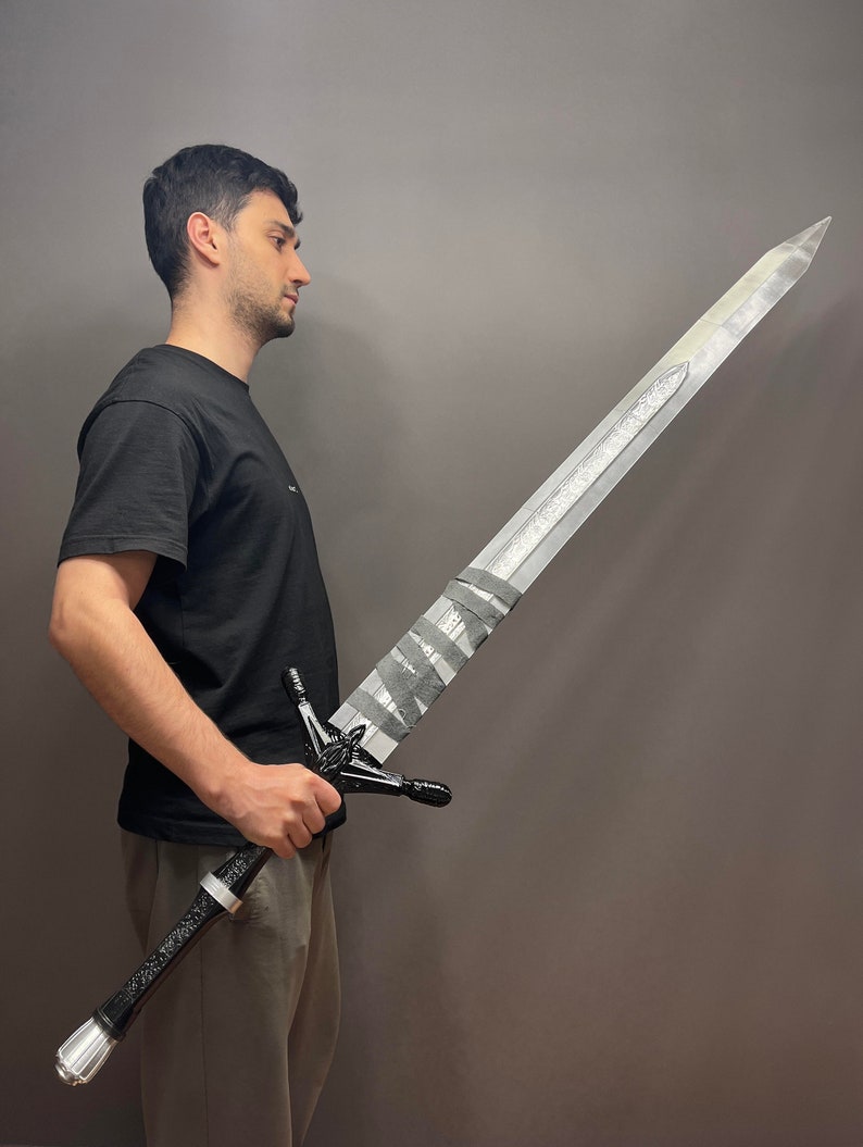 Puede incluir: Una persona sosteniendo una espada grande de color plateado con un mango negro y una empu&ntilde;adura en blanco y negro. La espada tiene un dise&ntilde;o decorativo en la hoja y una punta puntiaguda.