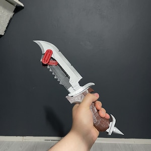 Könnte beinhalten: Ein silbernes und braunes Messer mit gezahnter Kante und rotem Verriegelungsmechanismus. Der Griff ist braun mit einem strukturierten Muster. Das Messer hat eine Spitze und eine gebogene Klinge. Das Messer wird in einer Hand gehalten.