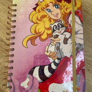 Könnte beinhalten: Ein rosa Notizbuch mit Spiralbindung und einem goldenen Lesezeichenband. Der Einband zeigt eine Comic-Illustration einer blonden Frau mit einer roten Schleife im Haar, die einen Waschbären hält.