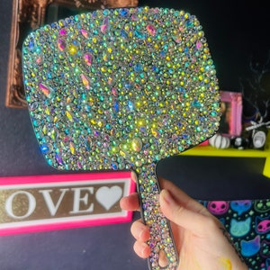 May include: A black hand mirror with a handle covered in iridescent rhinestones. The mirror is shaped like a rectangle with rounded corners.