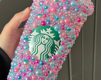 May include: A clear plastic tumbler covered in iridescent pink, blue, and white pearls with a green Starbucks logo.