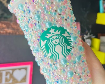 Op de afbeelding: Een doorzichtige plastic beker met een groen Starbucks-logo is bedekt met parels in pastelblauw, roze en wit.