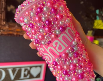 Op de afbeelding: Een roze plastic beker bedekt met roze en witte parels met de naam "Shan" erop.