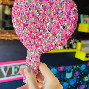 Può includere: Uno specchio da mano a forma di cuore ricoperto di strass rosa e bianchi iridescenti. Lo specchio ha un manico ed è tenuto in una mano.